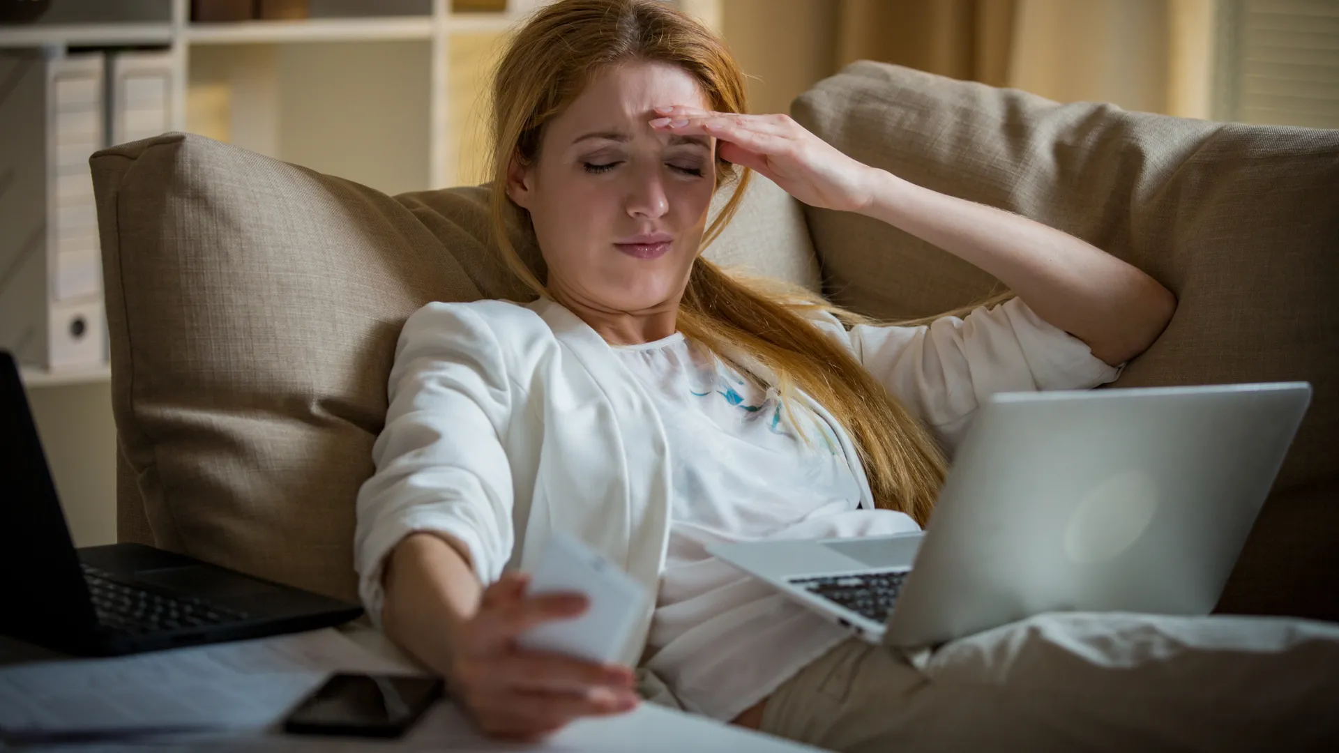 Nữ doanh nhân mệt mỏi ngồi trên ghế sofa cùng hai điện thoại và laptop, tay ôm thái dương
